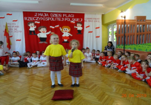 Na tle biało-czerwonej dekoracji stoją 2 dziewczynki w zółtych podkoszulkach i czerwonych spódnicach w kratę. Dziewczynki recytują wierszyk.